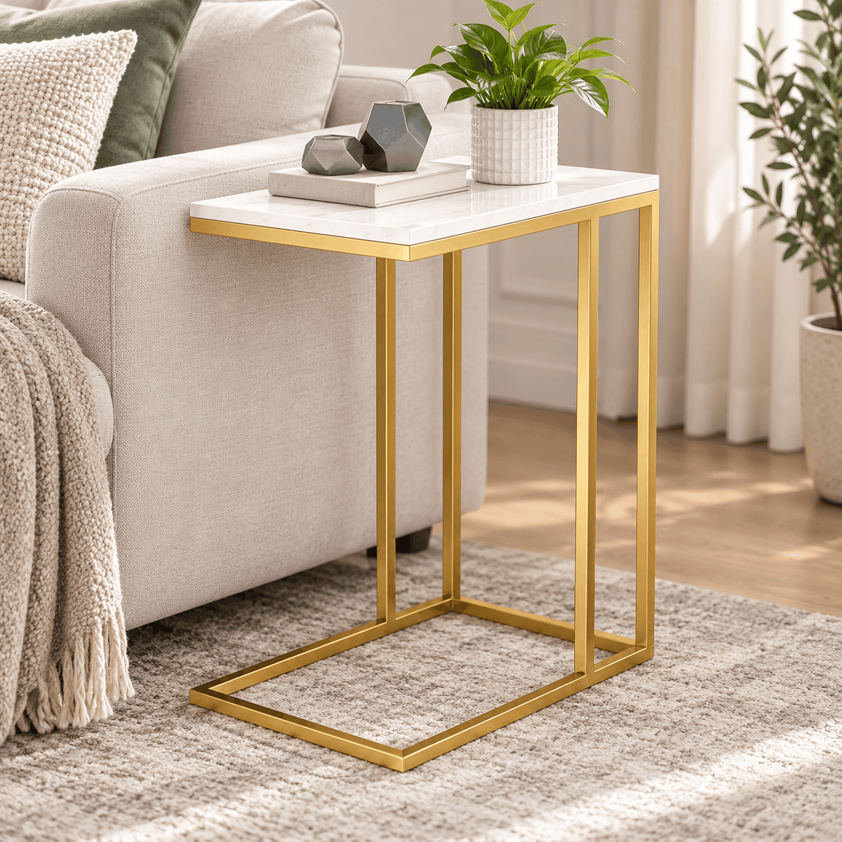 Gold frame side table with a white marble look top holding a plant, a book and two grey decor pieces beside a sofa arm