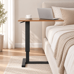 Height adjustable table with a wood look top and black frame beside a bed holding a laptop and a coffee cup