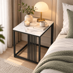 Square white marble look side table with a black slatted frame beside a bed holding a lamp, a vase, a candle and a book