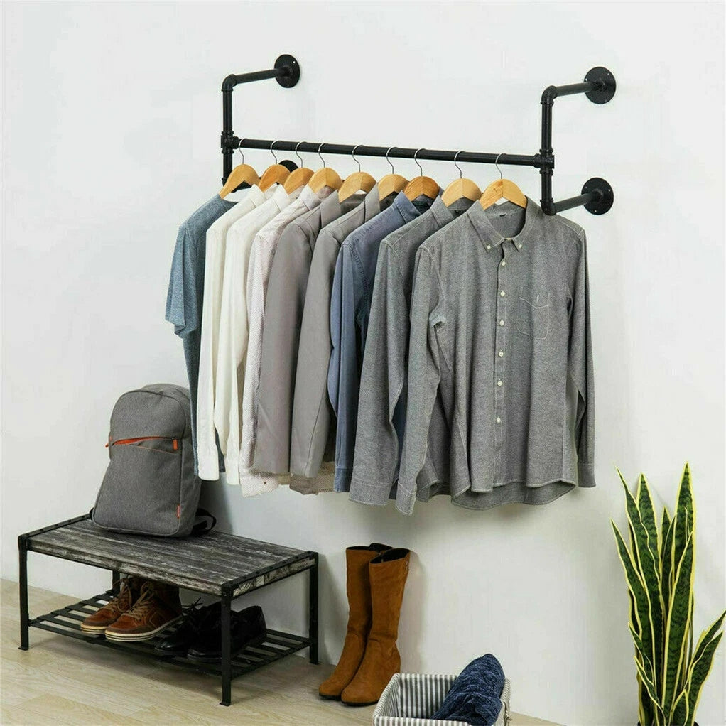 Black industrial pipe wall mounted clothes rack with shirts on wooden hangers above a shoe bench, backpack and boots