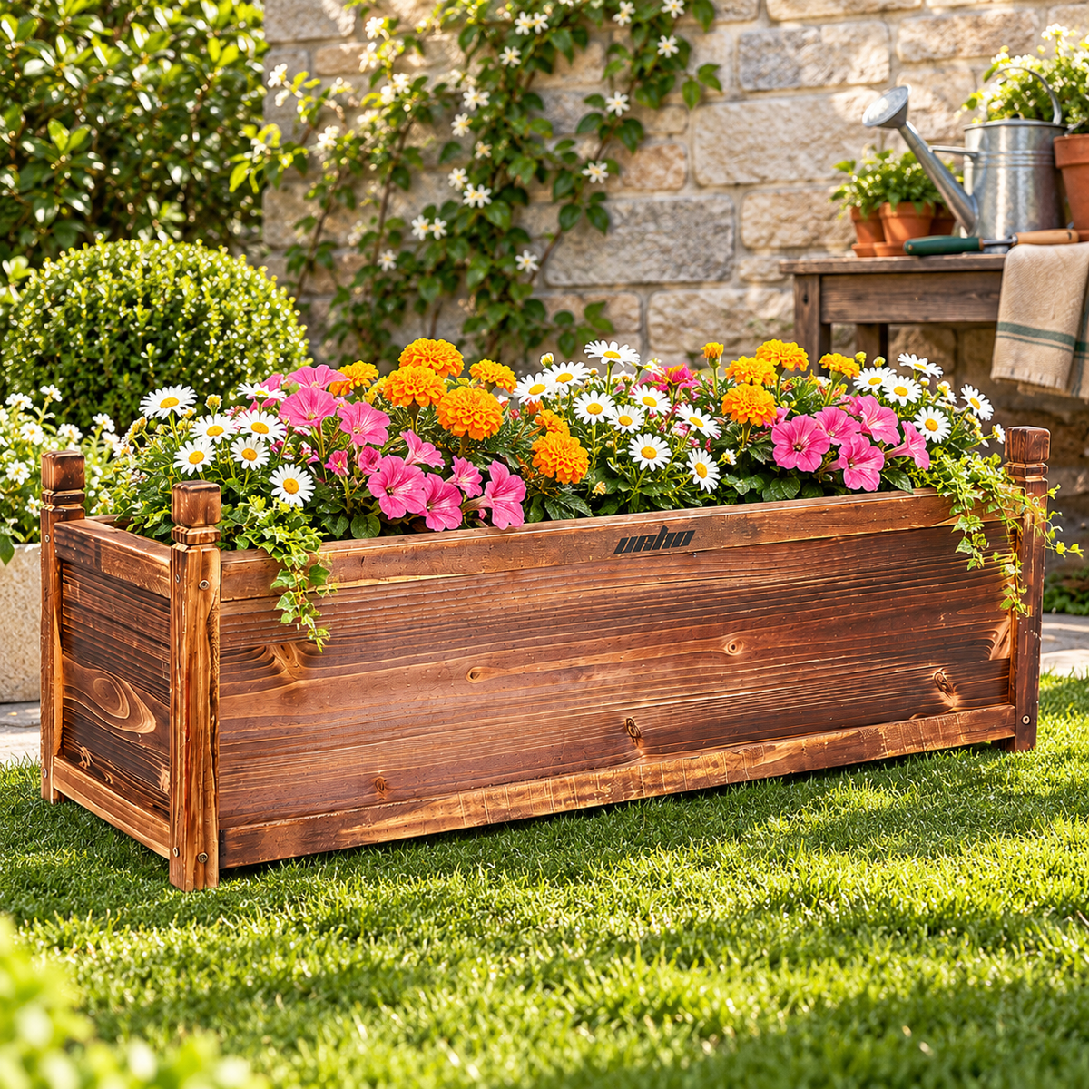 Long wooden planter box with marigolds, petunias and daisies on a lawn beside a stone wall and potting table